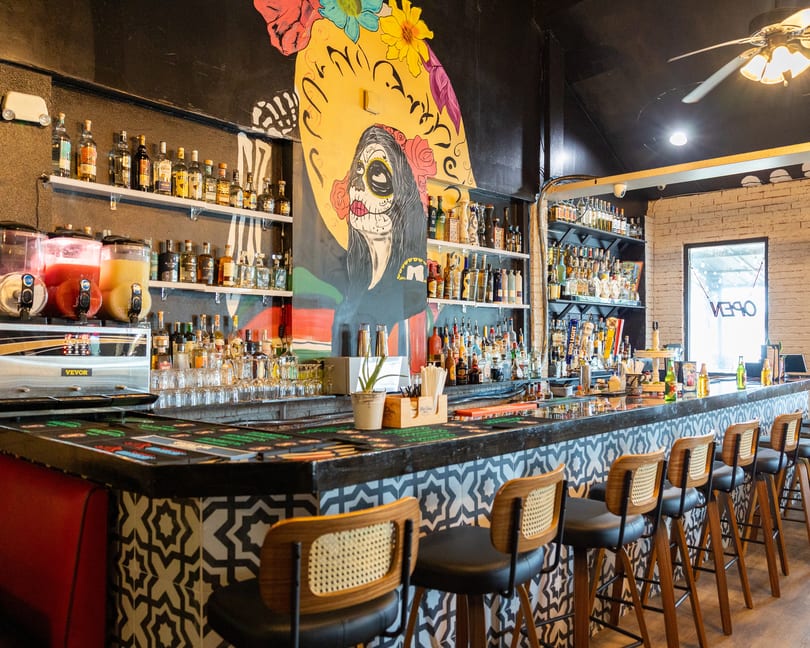 A vibrant bar with a Day of the Dead mural, liquor bottles, margarita machines, and bar stools.