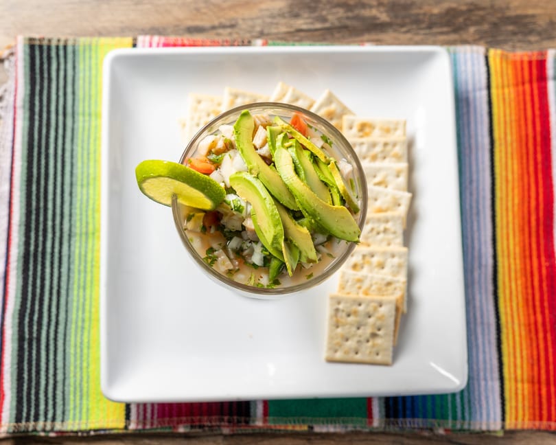 Ceviche with avocado slices and a lime wedge, served with crackers on a white plate and colorful striped mat.