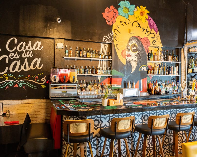 A vibrant Mexican restaurant bar with a colorful Day of the Dead mural and rows of liquor bottles.