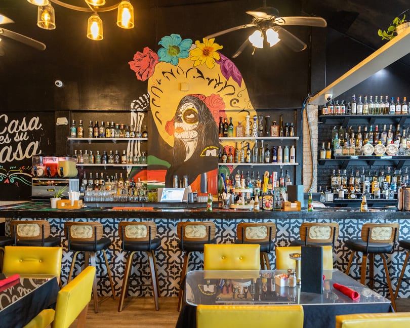 A vibrant bar with a large Day of the Dead mural, liquor bottles, and yellow chairs.