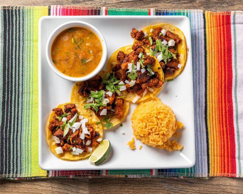 Four tacos al pastor with rice, salsa, and a lime wedge on a white plate.