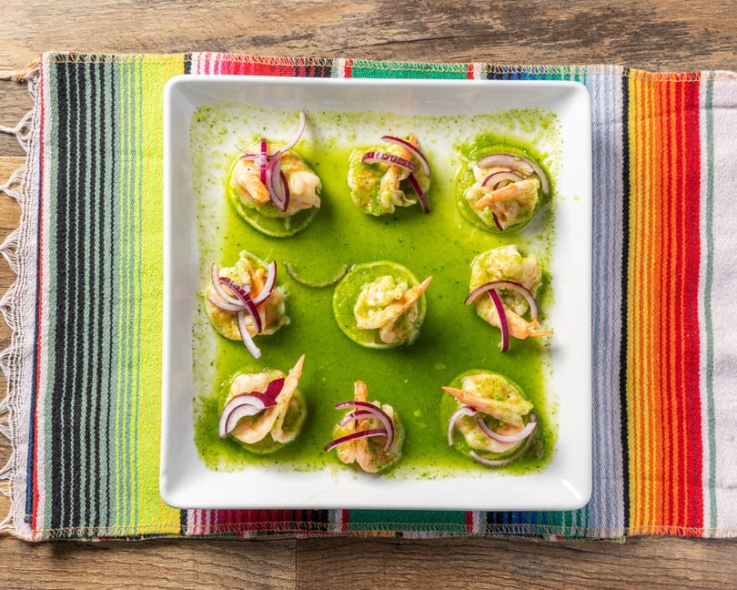 Nine shrimp aguachile bites with red onion on lime slices in green sauce on a white plate.