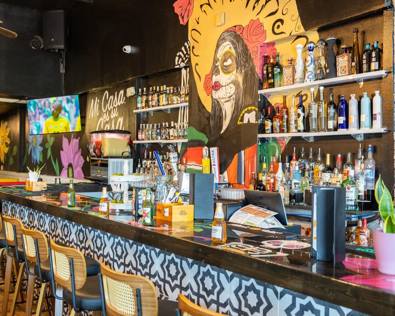 A vibrant bar with a Dia de los Muertos mural, stocked shelves of liquor, and a TV showing sports.