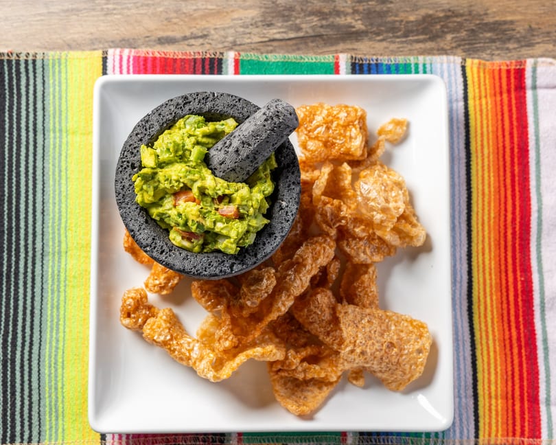 Guacamole in a molcajete with chicharrones on a white plate, on a colorful striped placemat.