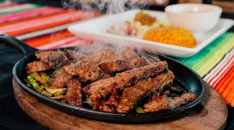 Sizzling steak fajitas with peppers and onions in a cast iron skillet, with sides in the background.