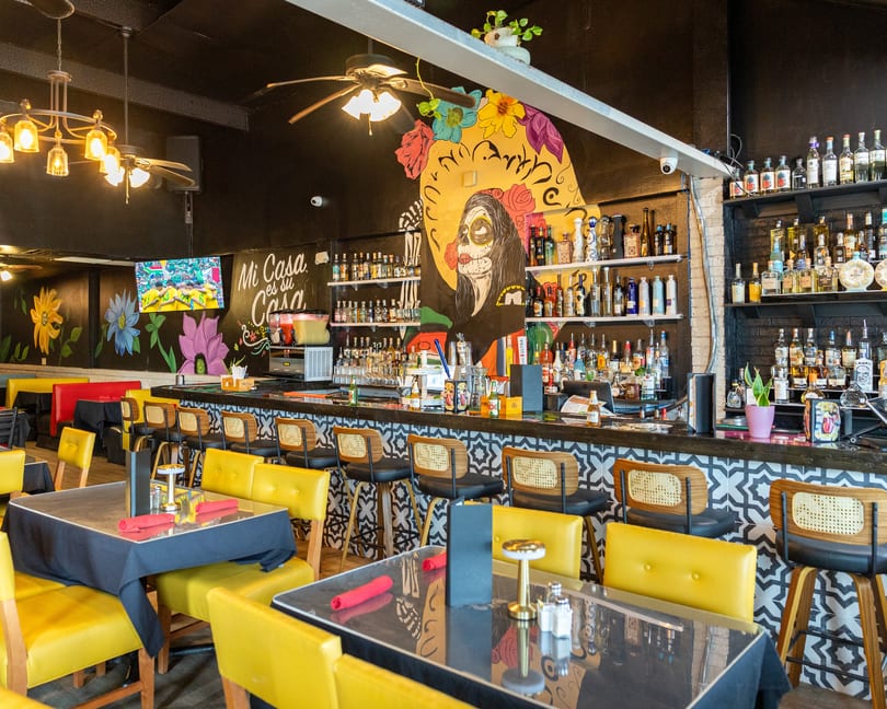 A vibrant restaurant interior with a bar, yellow chairs, and a large colorful mural of a woman.