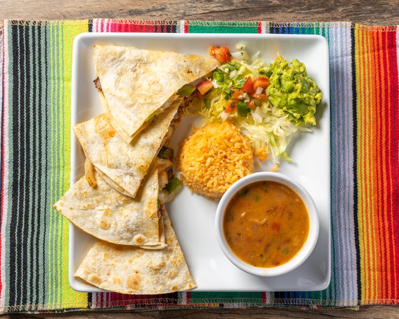 Quesadillas with rice, guacamole, salsa, and bean soup on a white plate over a colorful striped mat.