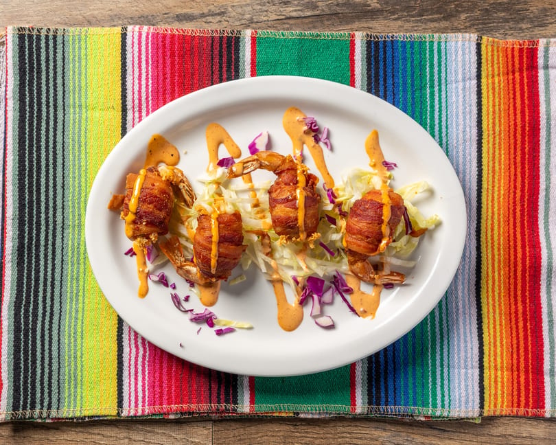 Four bacon-wrapped shrimp with sauce and shredded lettuce on a white plate, on a colorful striped placemat.