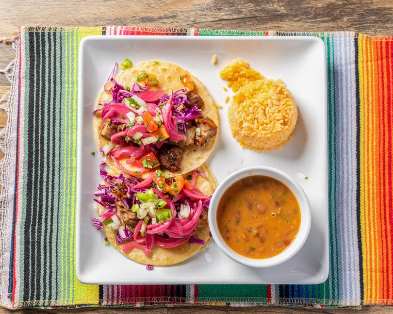 Two tacos with meat, red onions, and cabbage, served with rice and beans on a colorful placemat.