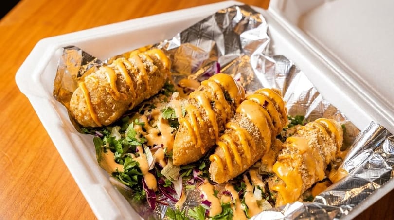 Four crispy fried taquitos drizzled with orange sauce and sprinkled with cheese, served over salad in a to-go container.