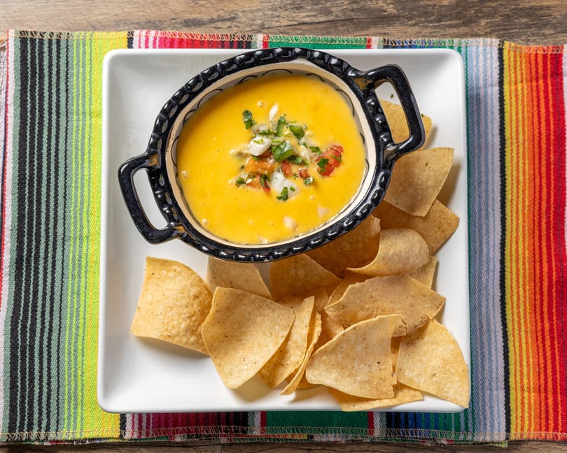 Queso dip with pico de gallo and tortilla chips on a white plate, set on a colorful striped placemat.