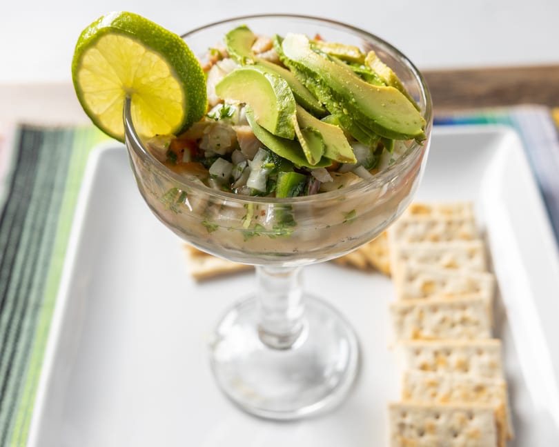 Ceviche in a glass, garnished with avocado slices and a lime wedge, served with crackers.