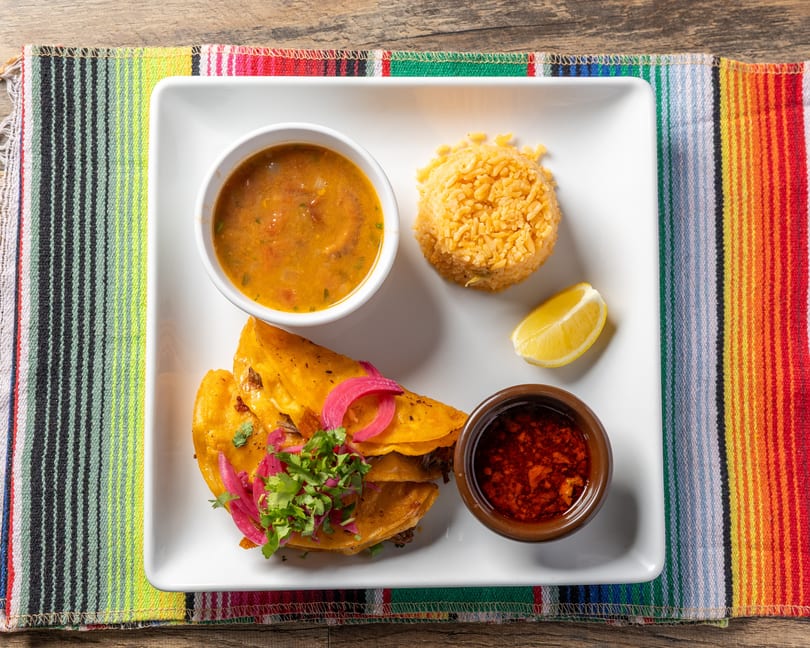 Two birria tacos with pickled onions and cilantro, rice, consommé, chili oil, and a lemon wedge.