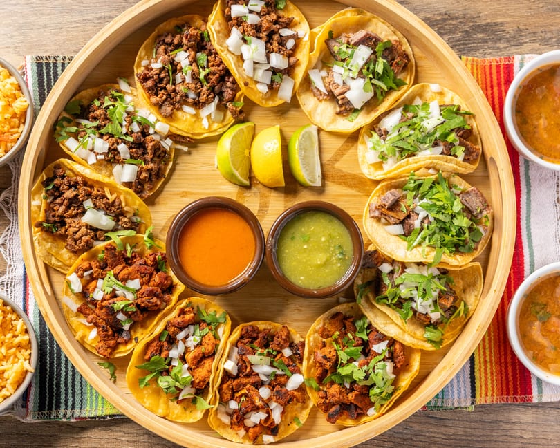 A wooden tray filled with various tacos, two bowls of rice, and two bowls of soup.