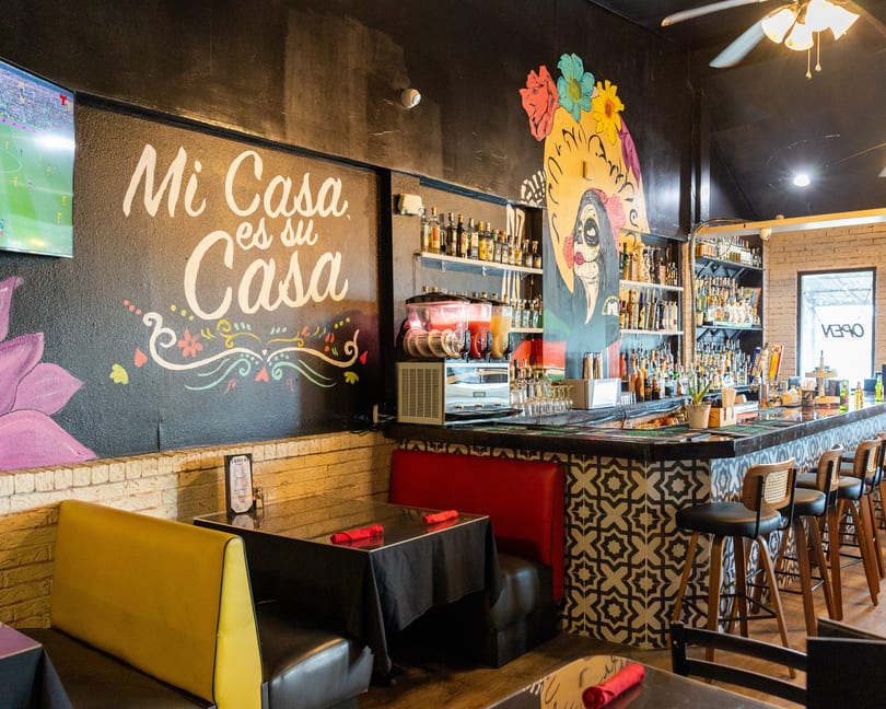 Vibrant Mexican restaurant interior with a bar, colorful mural, "Mi Casa es Su Casa" sign, and booths.